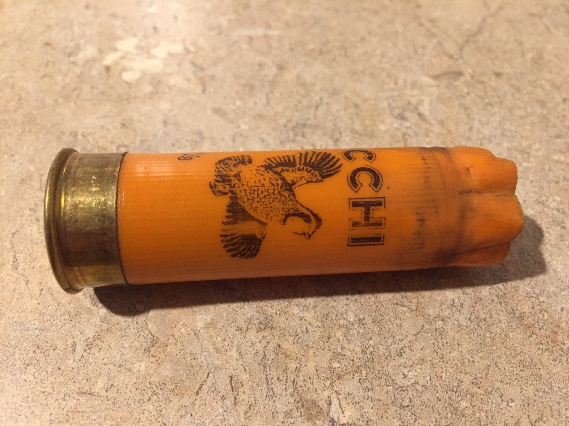 One horizontal orange 12 gauge shotshell with printed pheasant graphic lying on a speckled stone surface.