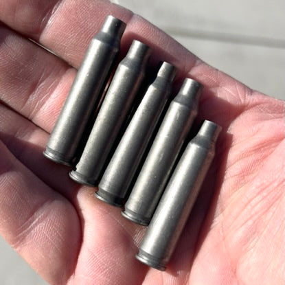 A hand holds five once-fired gray steel cartridges lying side by side across the palm.