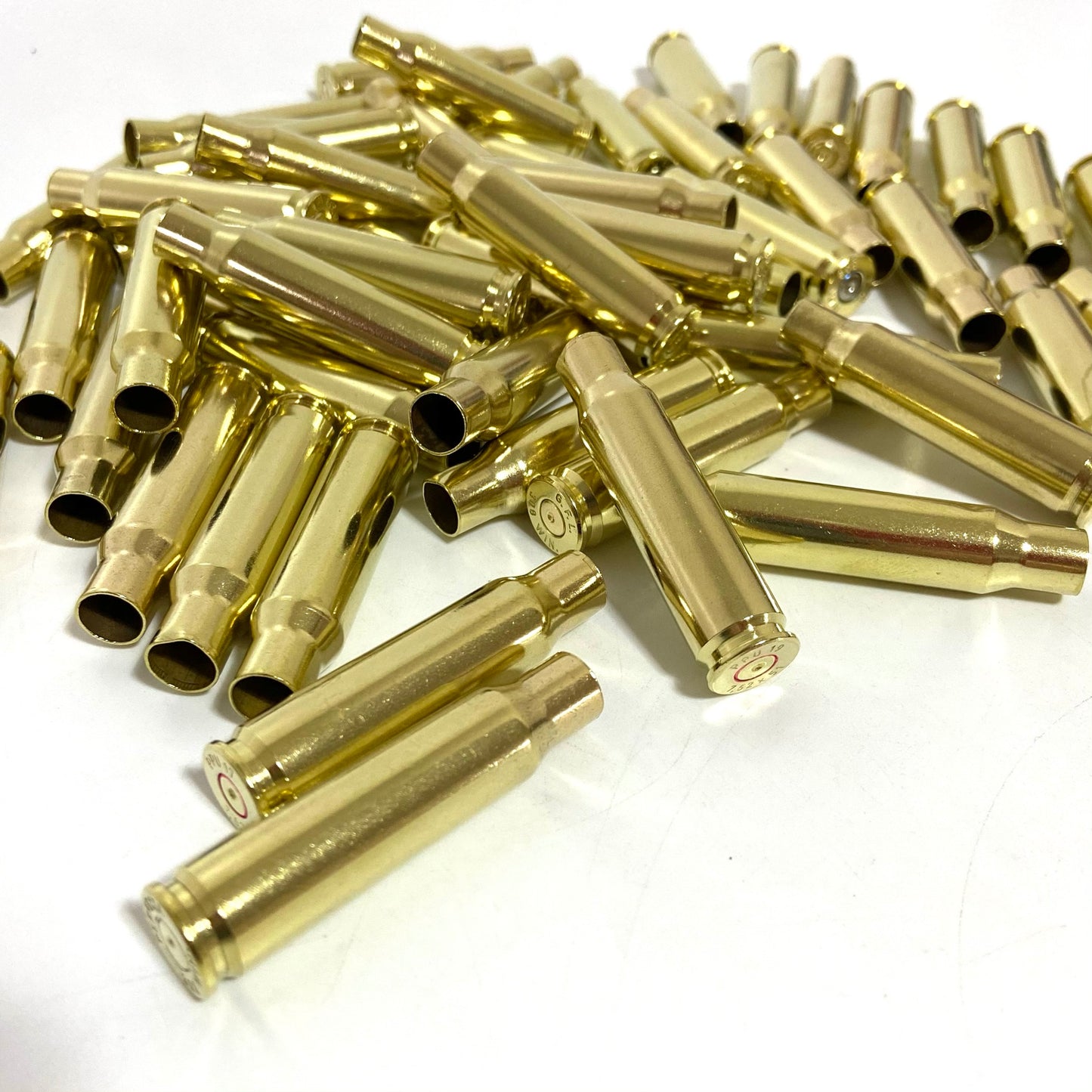 Bulk mound of gold 308 WIN brass cartridges on a white surface with two foreground cases showing base rims.