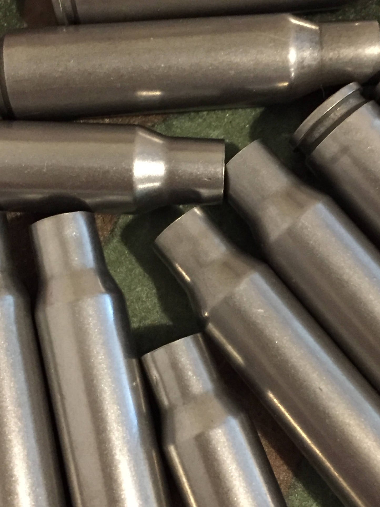 Closeup of dark gray steel 308 Winchester rifle casings lying on a green surface, showing bottlenecked shoulders