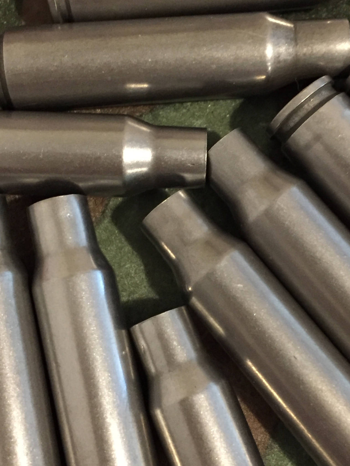 Closeup of dark gray steel 308 Winchester rifle casings lying on a green surface, showing bottlenecked shoulders