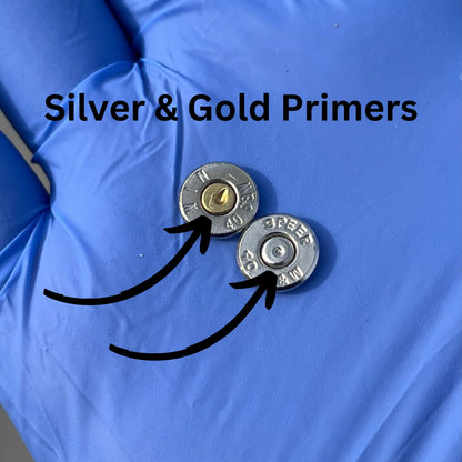 Two polished 40 S&W nickel bullet slices resting on a blue glove, featuring 'Silver & Gold Primers' text and black arrows pointing to a gold-colored primer on the left and a silver-colored primer on the right. 