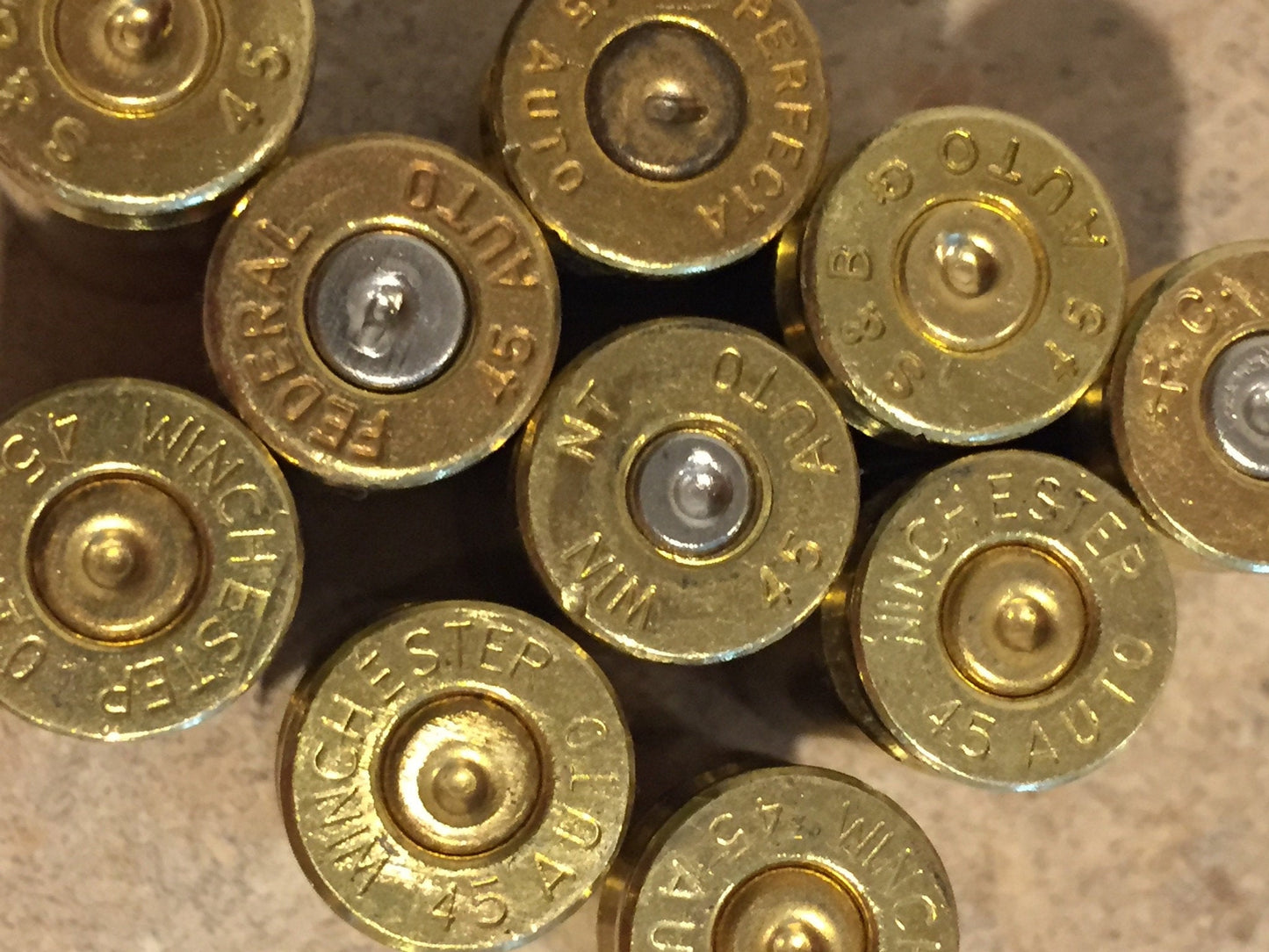 Close-up of .45 ACP brass casing headstamps on a stone surface, clearly showing "Winchester 45 Auto," "Federal 45 Auto," and "Perfecta 45 Auto" markings with mixed silver and gold-toned primers