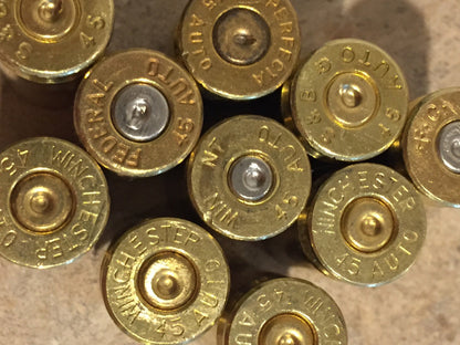 Close-up of .45 ACP brass casing headstamps on a stone surface, clearly showing "Winchester 45 Auto," "Federal 45 Auto," and "Perfecta 45 Auto" markings with mixed silver and gold-toned primers