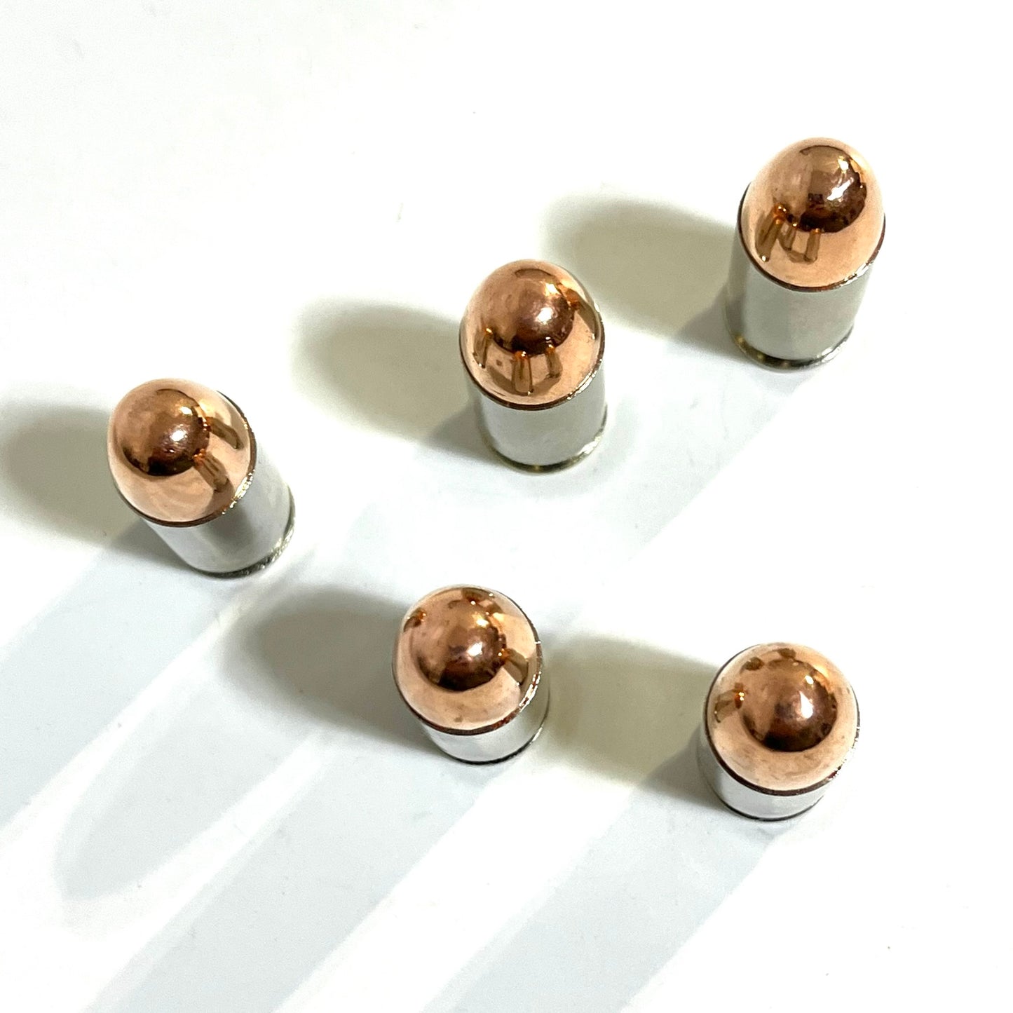 An angled overhead view of five nickel 45 ACP snap caps standing on a white background with long shadows.