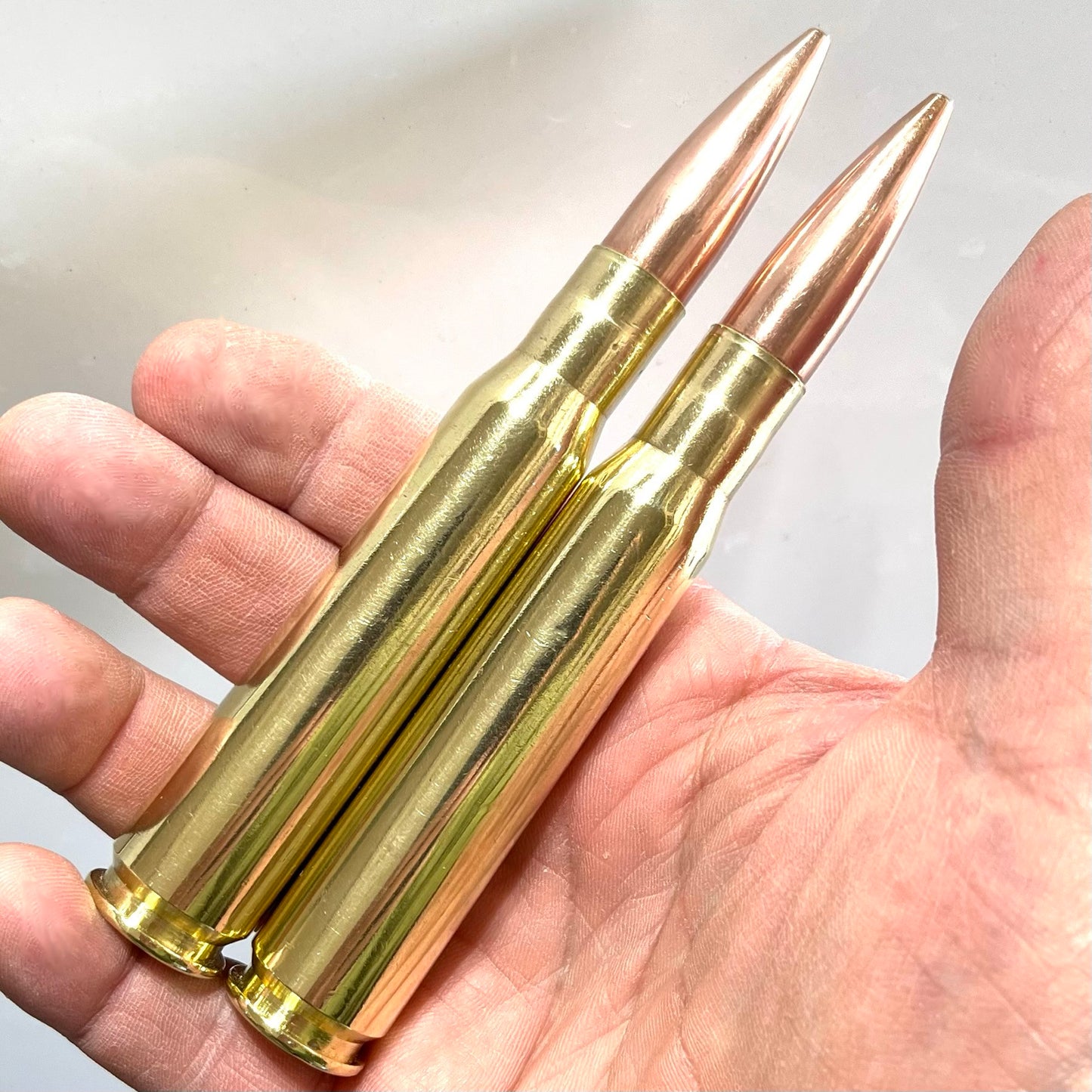 Two shiny brass 50 BMG paperweight rounds with copper bullets resting in an open palm of a bare hand.
