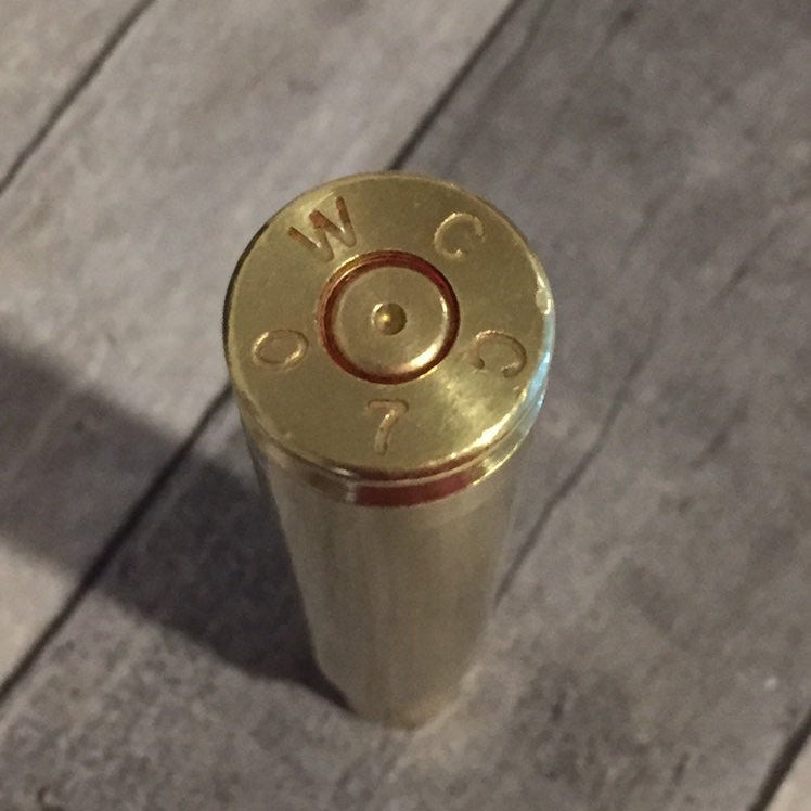 Base of a polished .50 BMG brass casing stamped WCC, with a dimpled spent primer, standing on gray wood