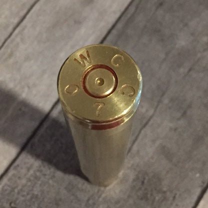 Base of a polished .50 BMG brass casing stamped WCC, with a dimpled spent primer, standing on gray wood
