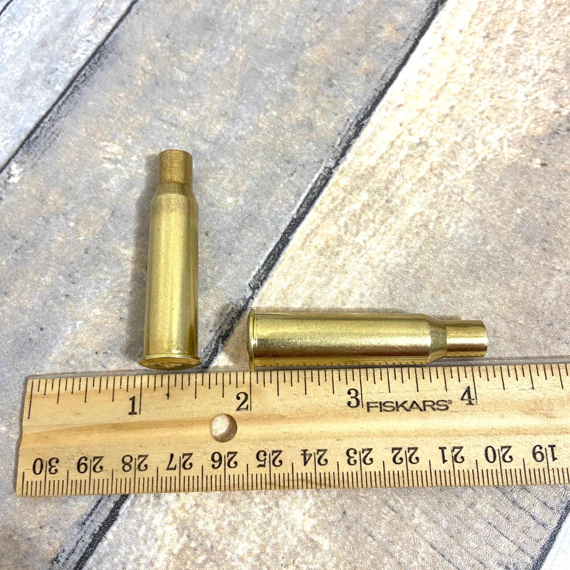 Two polished 7.62x54R empty brass casings shown next to a wooden ruler by Empty Shotgun Shells for size reference.
