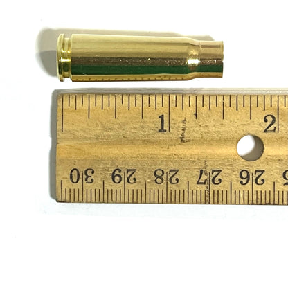 Once-fired brass shell with a green-tinted band around its body, resting above a wooden ruler for scale.