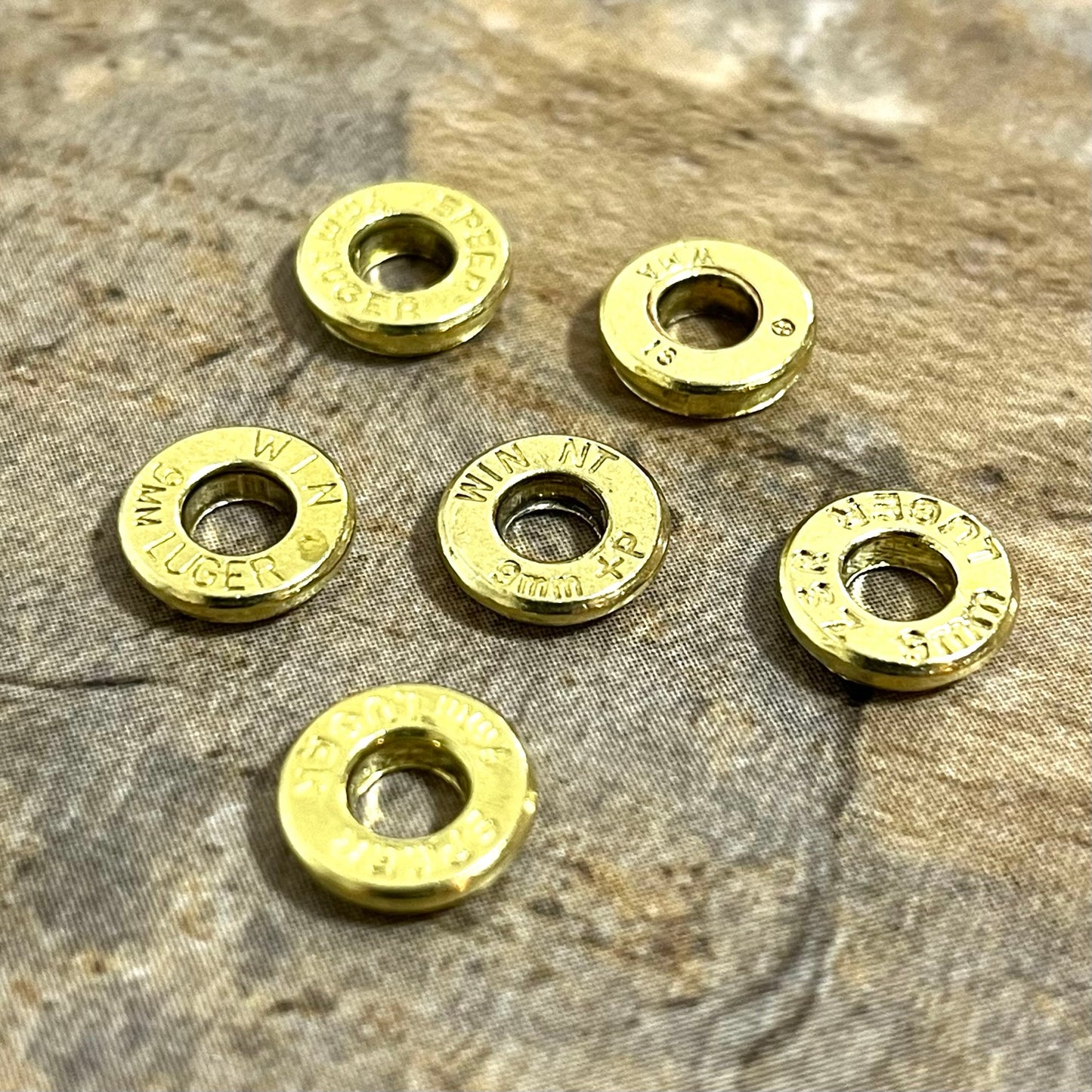 Six individual brass slices with primer removed rest on a stone surface, each stamped with different headstamps.
