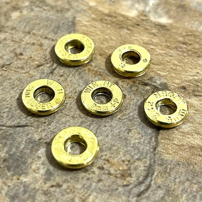 Six individual brass slices with primer removed rest on a stone surface, each stamped with different headstamps.
