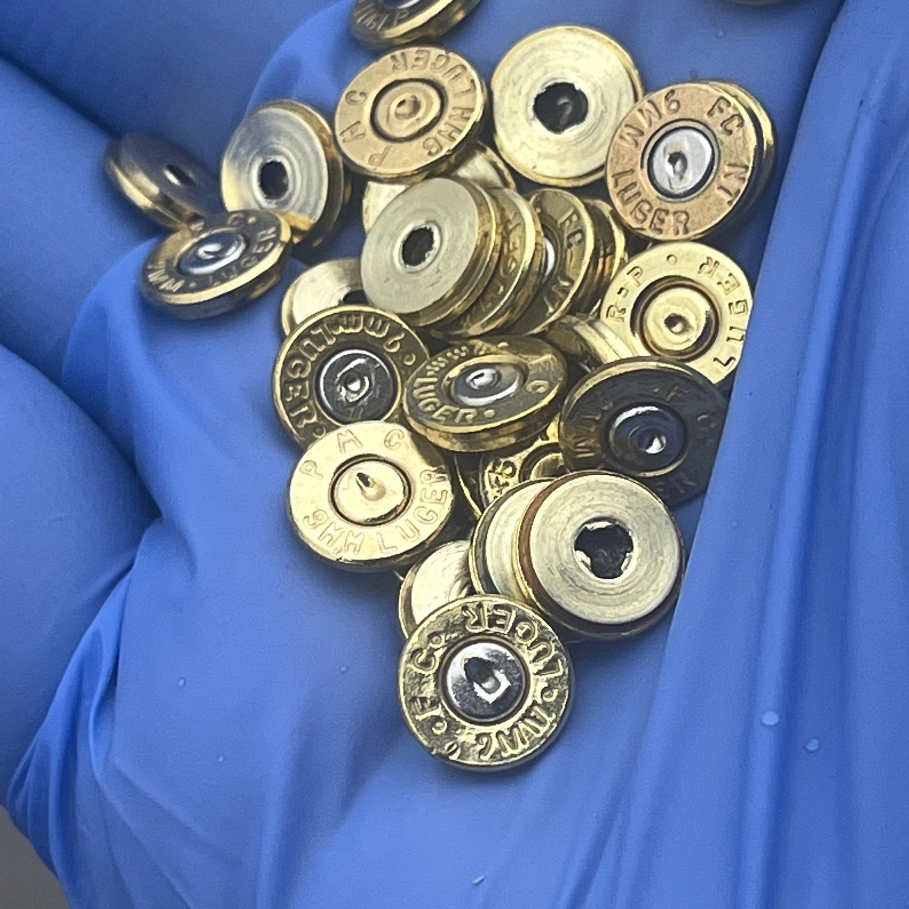 A collection of polished brass 9mm bullet slices held in gloved hands, displaying different headstamps.
