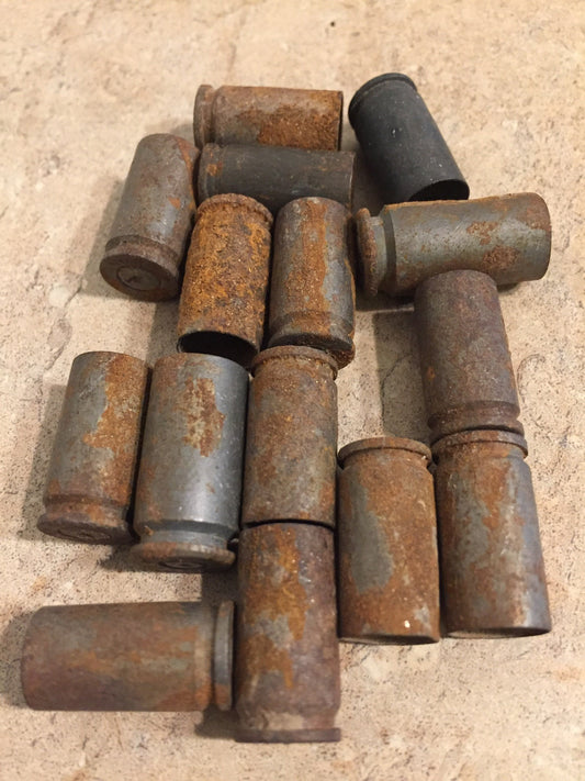 Pile of 15 rusted 9mm brass shell casings on tan tile surface, heavy orange and gray patina, spent primers visible