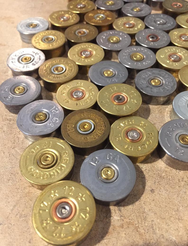 An angled top-down view of mixed gold and silver 12 gauge shotgun shell headstamps with spent primers on a textured surface.
