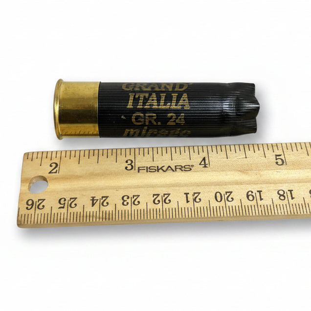 Black Mirage Grand Italia shotgun hull lying beside a wooden ruler for length comparison.