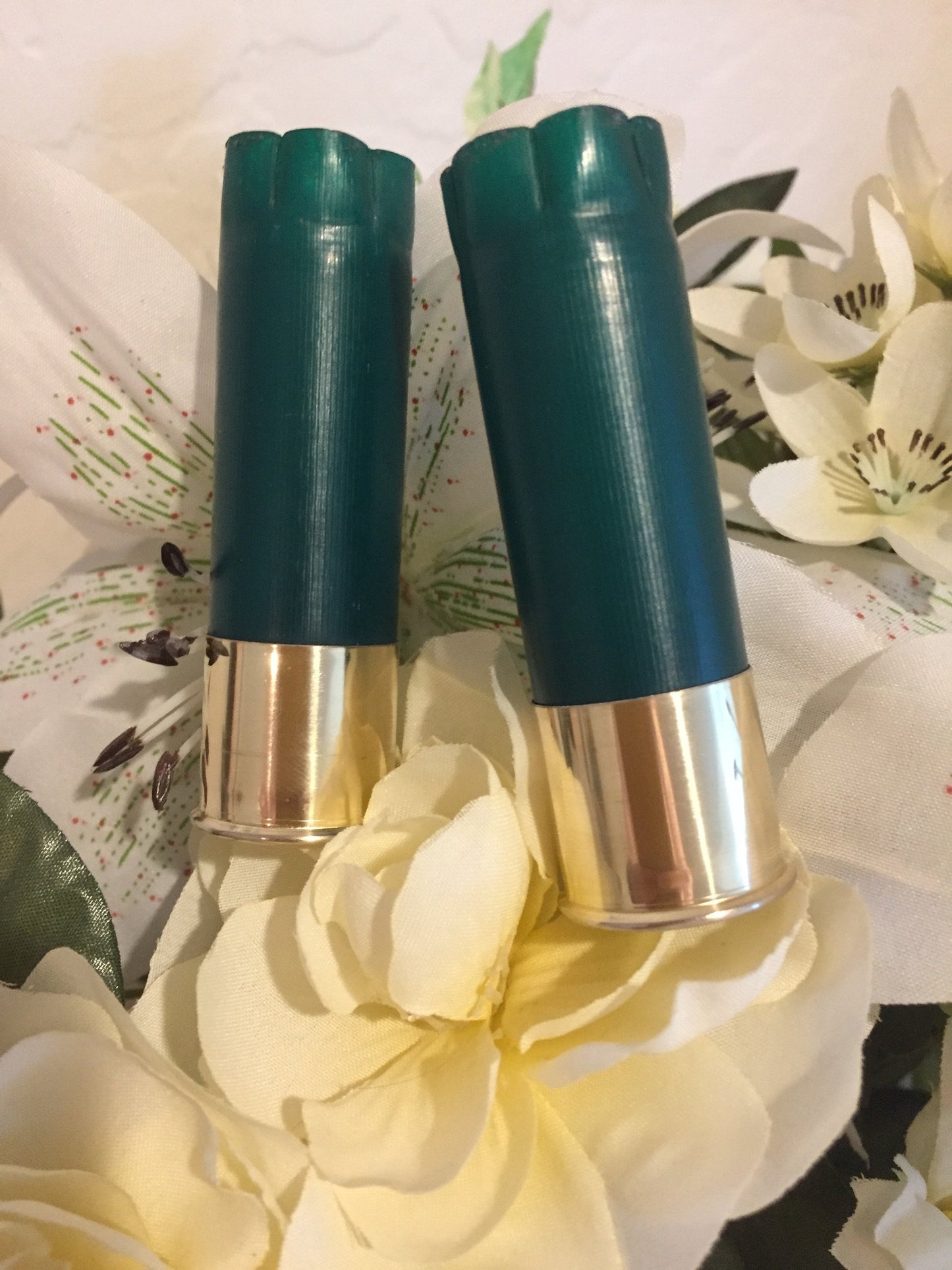 Two empty green shotgun shells standing upright surrounded by white silk flowers.