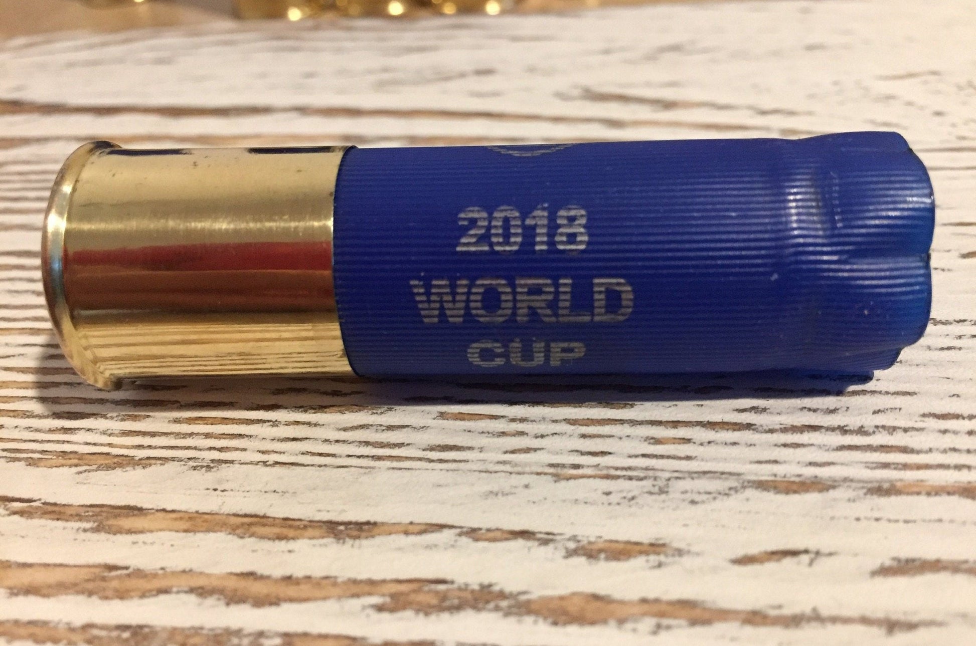 Closeup of a blue ‘2018 World Cup’ shotgun shell with a gold brass base lying on a wood-plank background