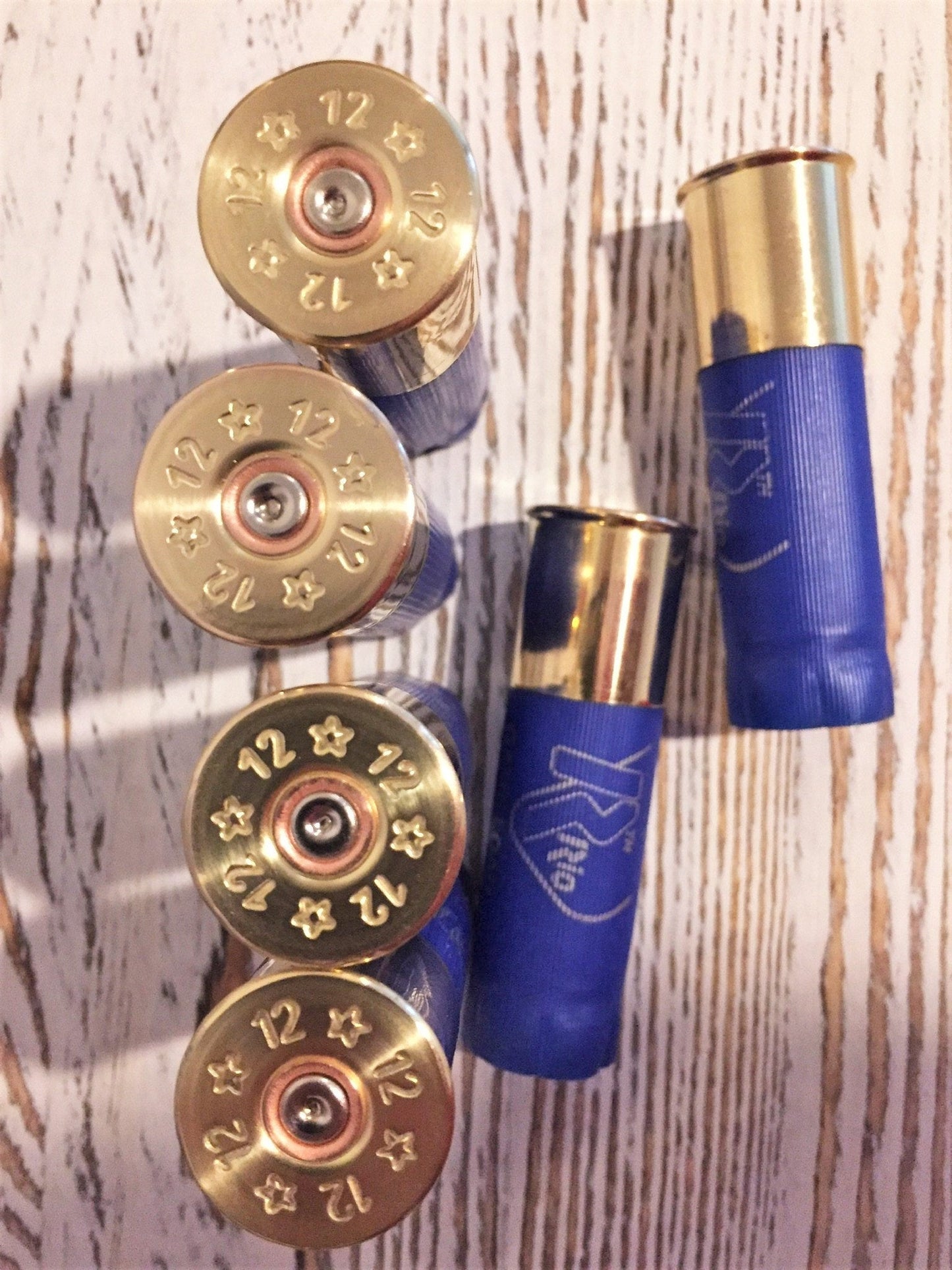 Six blue shotgun shells with gold brass bases arranged on a wood-plank background, showing ‘12’ star stampings on four bases and printed ‘Rio’ text on two hulls