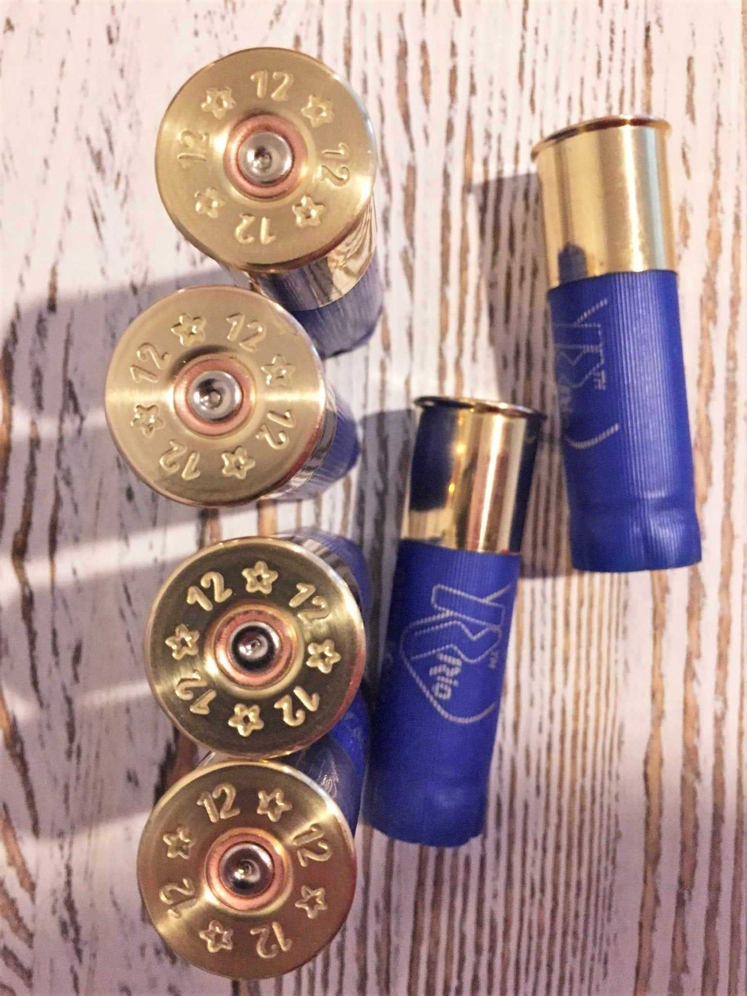 Six blue shotgun shells with gold brass bases arranged on a wood-plank background, showing ‘12’ star stampings on four bases and printed ‘Rio’ text on two hulls