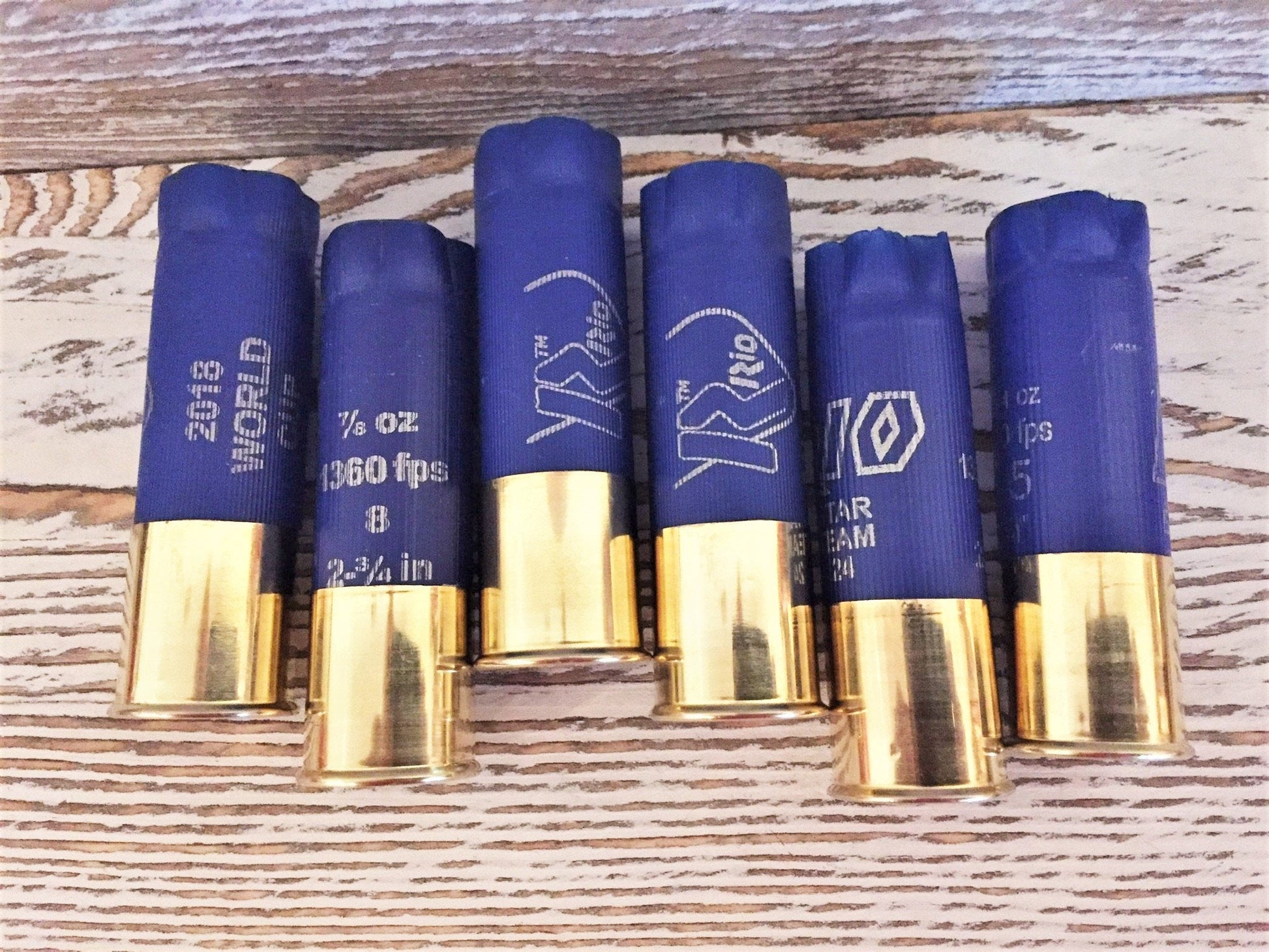 Six blue shotgun shells with gold brass bases standing upright on a wood-plank background, showing various printed brand markings including ‘Rio’ and ‘2018 World Cup’