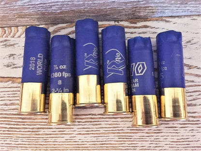 Six blue shotgun shells with gold brass bases standing upright on a wood-plank background, showing various printed brand markings including ‘Rio’ and ‘2018 World Cup’