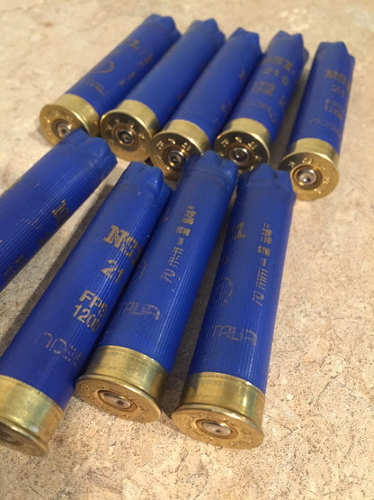 Nine blue 28 gauge shotgun shells lying diagonally with brass crimped ends, showing “Noble” and “Italia” branding