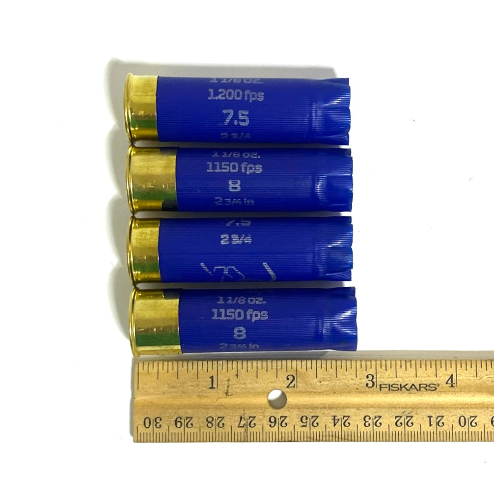 Four blue Rio 12 gauge shotgun hulls aligned horizontally next to a wooden Fiskars ruler showing length.