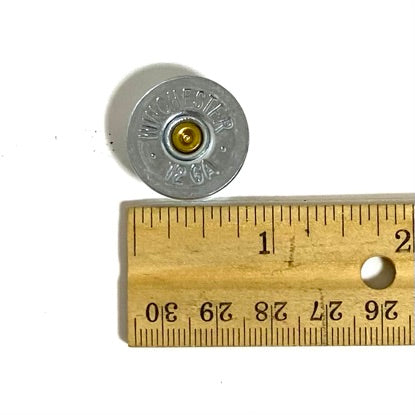 Silver 12 gauge Winchester shotgun shell bottom stamped '12 GA', shown beside a wooden ruler for scale.