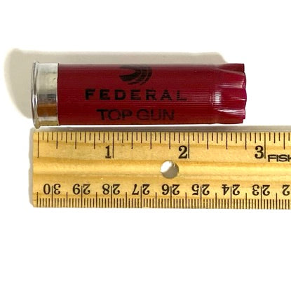 One burgundy shotgun casing placed horizontally above a wooden ruler showing a length of approximately two point seventy-five inches.