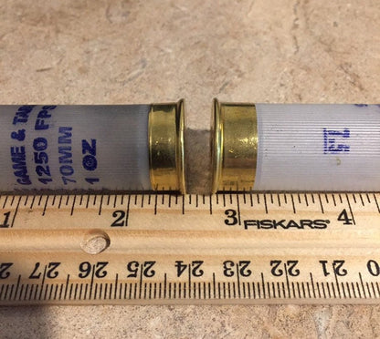 Two translucent white Fiocchi 12 gauge shotgun shells lying next to a wooden ruler showing their length