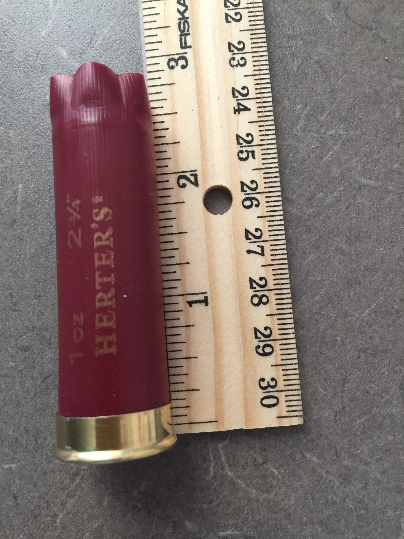 Single burgundy Herter’s 12 gauge shotgun shell measured against a ruler, showing a length of about 2.75 inches