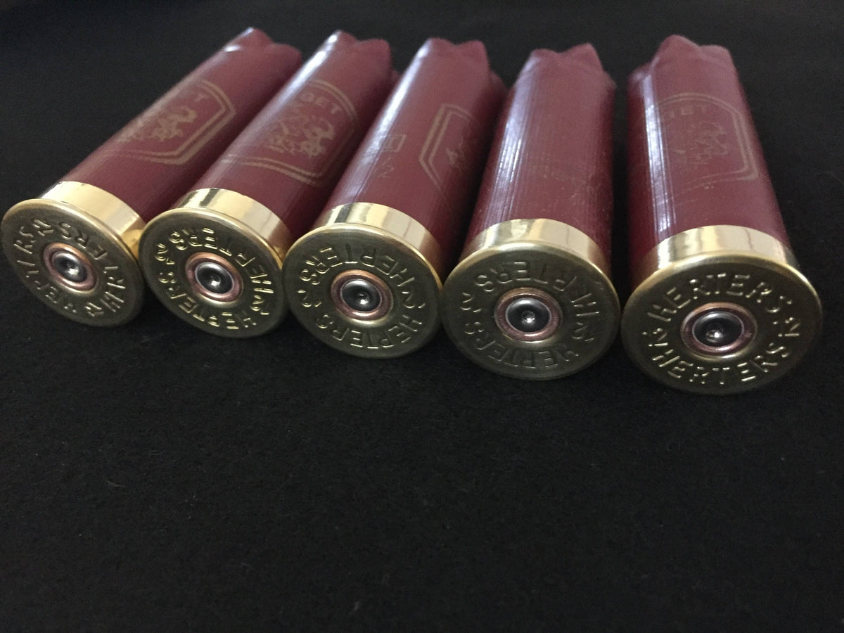 Five gold Herter’s 12 gauge shotgun shell bases viewed at an angle on a black background, showing burgundy hulls and stamped ‘Herters 12’ markings