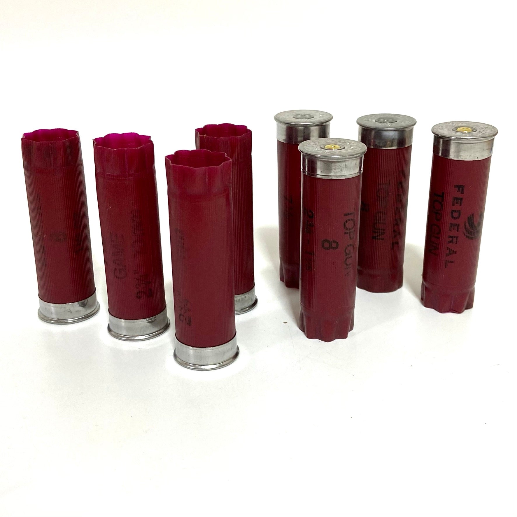 Seven empty dark red shotgun shells standing upright in two rows on a white surface.
