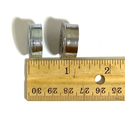 Two silver 12 gauge shotgun shell end caps standing on edge, showing their side profile next to a ruler.