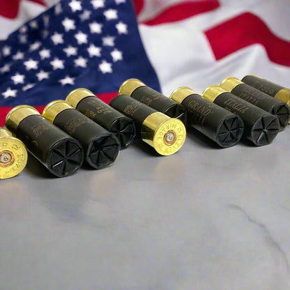 Six black spent hulls with gold brass bottoms arranged on a surface with an American flag in the background.