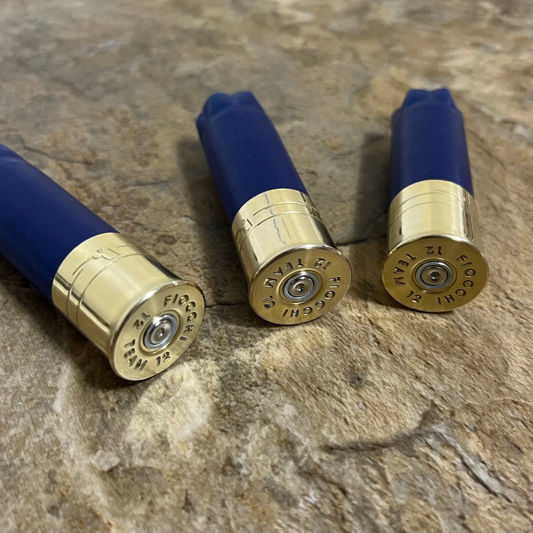 Three blue Fiocchi 12 gauge shotgun hulls on textured stone, showing gold high brass headstamps with readable Fiocchi text.