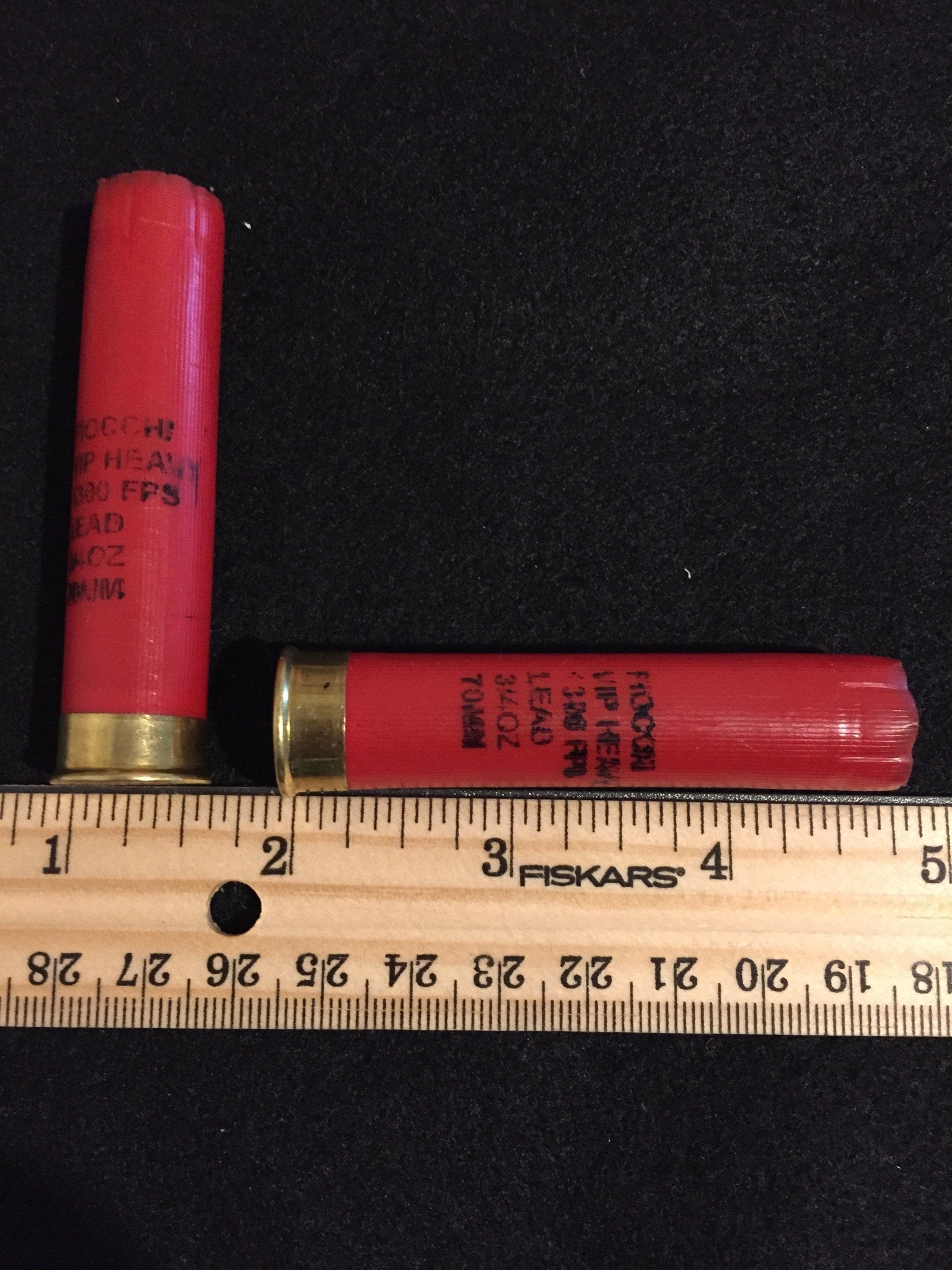 Two red Fiocchi 28 gauge shotgun shell hulls beside a wooden ruler, one marked 70mm on the hull