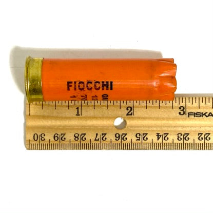 A single orange Fiocchi hull laid against a wooden ruler showing its length in inches.