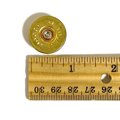 A gold 12 gauge shotgun shell headstamp positioned next to a wooden ruler for scale.
