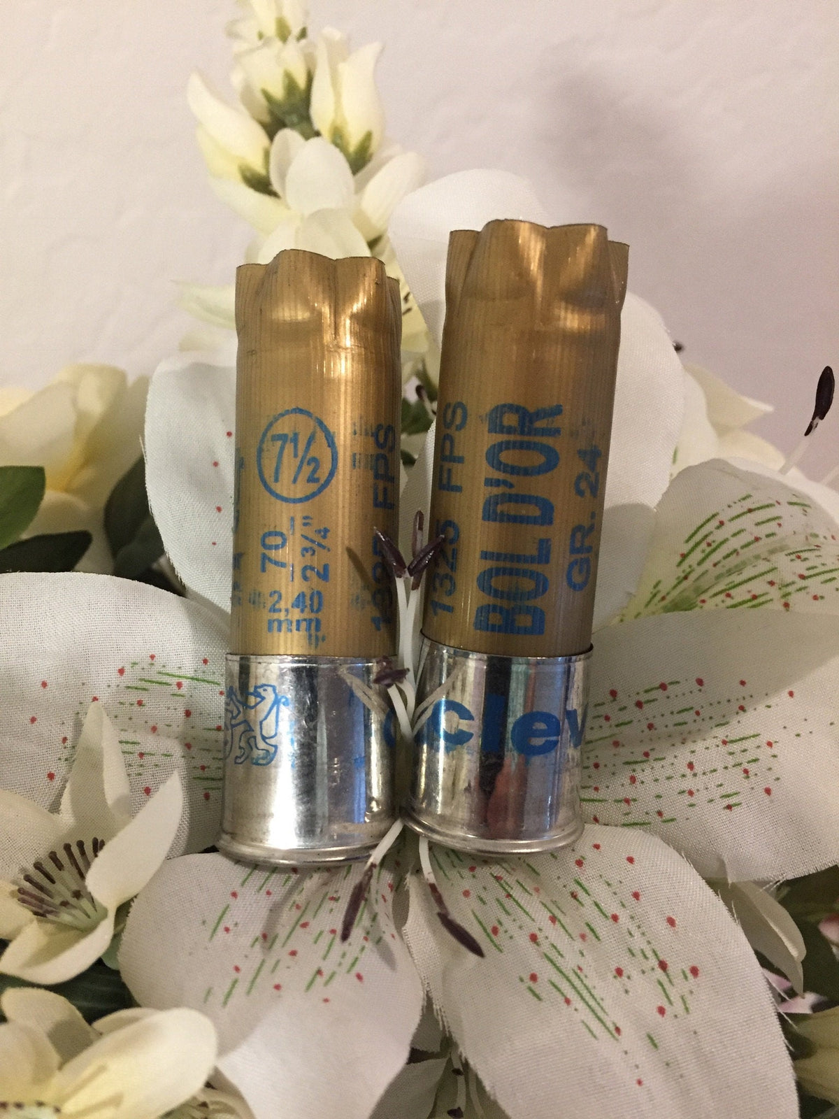 2 gold shotgun shells with silver bases standing upright printed "7½", "Bol D'Or", "1325 FPS", "GR.24" among white flowers