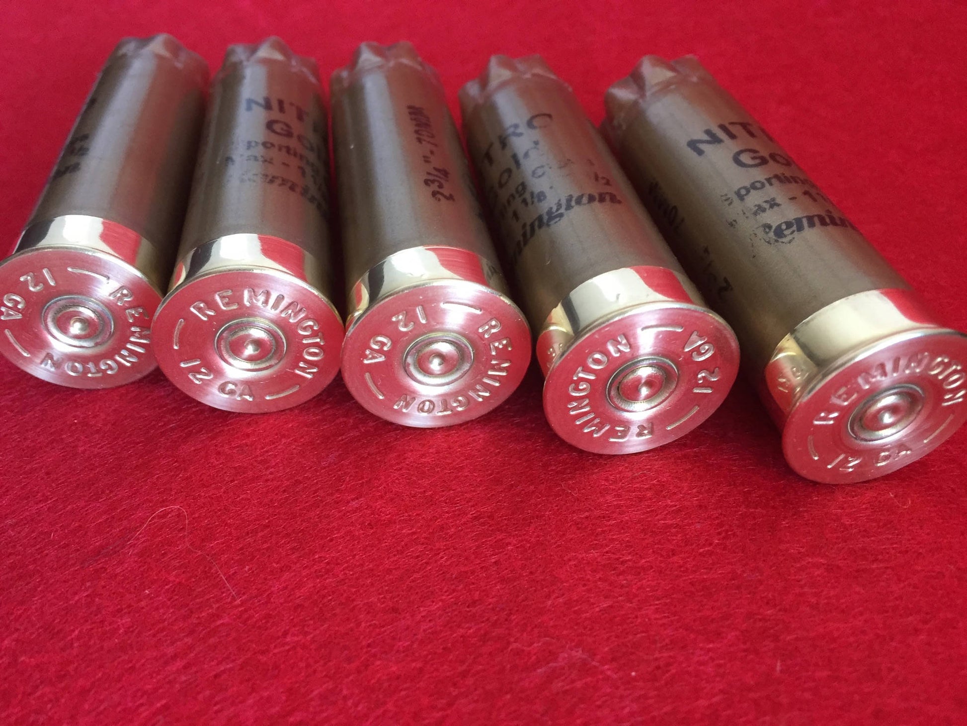 5 gold Remington Nitro shotgun shells lying in a row showing brass heads with REMINGTON 12 GA headstamp text on a red surface