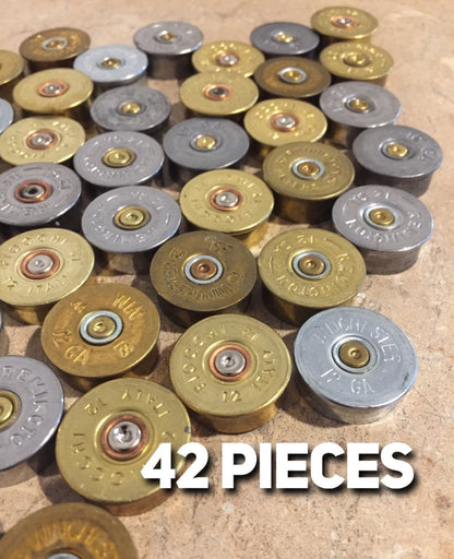 A collection of 42 gold and silver shotgun shell headstamps scattered on a neutral-colored surface with 42 PIECES" text overlay.

