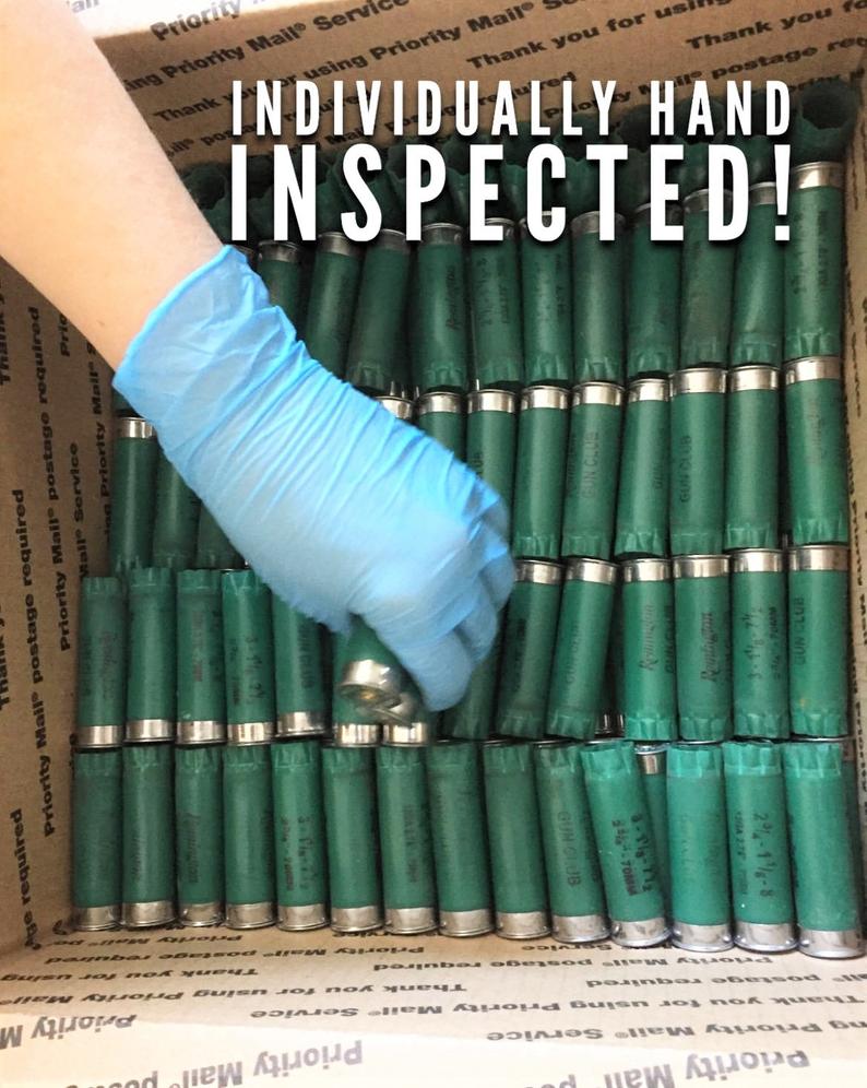 Rows of once-fired green 12ga Remington Gun Club hulls in a shipping box with a gloved hand and hand inspected text overlay