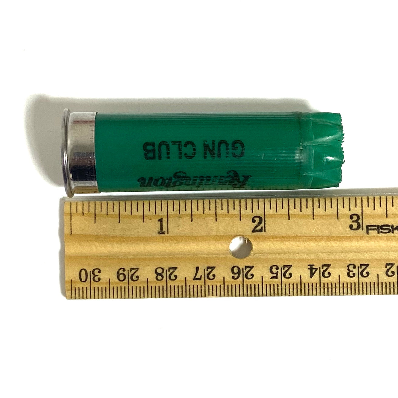 Used green 12ga Remington Gun Club cartridge with GUN CLUB text laid against a wooden ruler for size
