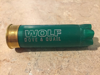 Single green shotgun shell with a brass base lying on a stone surface printed "Wolf Dove & Quail"