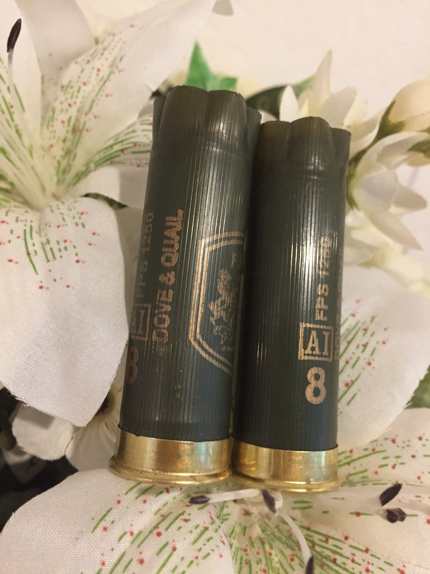 Two dark green “Dove & Quail” 12 gauge shotgun shells with brass bases resting on a white floral fabric background
