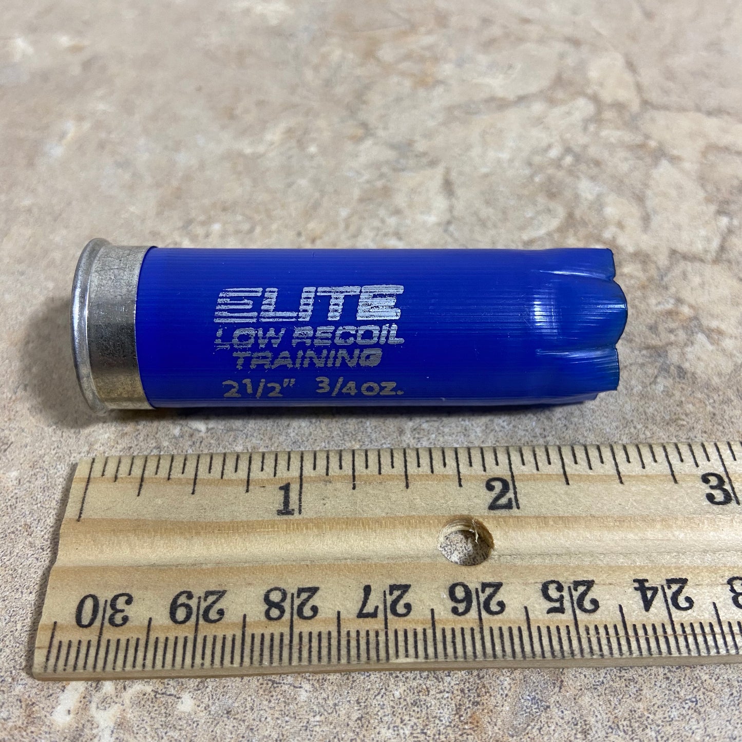 Horizontal view of a single navy blue Kent 12-gauge low recoil training empty shotgun shell lying next to a wooden ruler for size comparison showing its 2.50 inch length.

