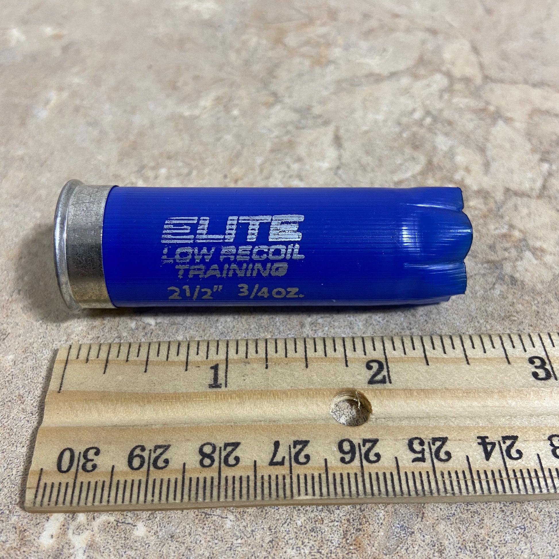 Horizontal view of a single navy blue Kent 12-gauge low recoil training empty shotgun shell lying next to a wooden ruler for size comparison showing its 2.50 inch length.
