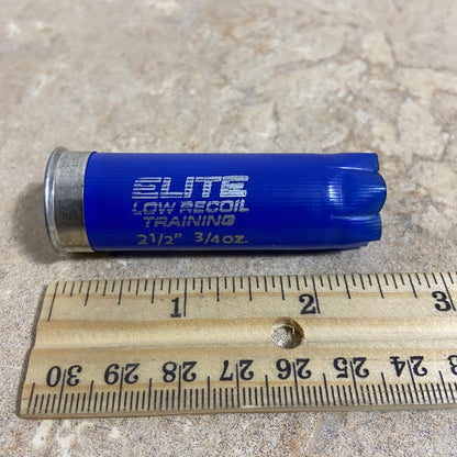 Horizontal view of a single navy blue Kent 12-gauge low recoil training empty shotgun shell lying next to a wooden ruler for size comparison showing its 2.50 inch length.
